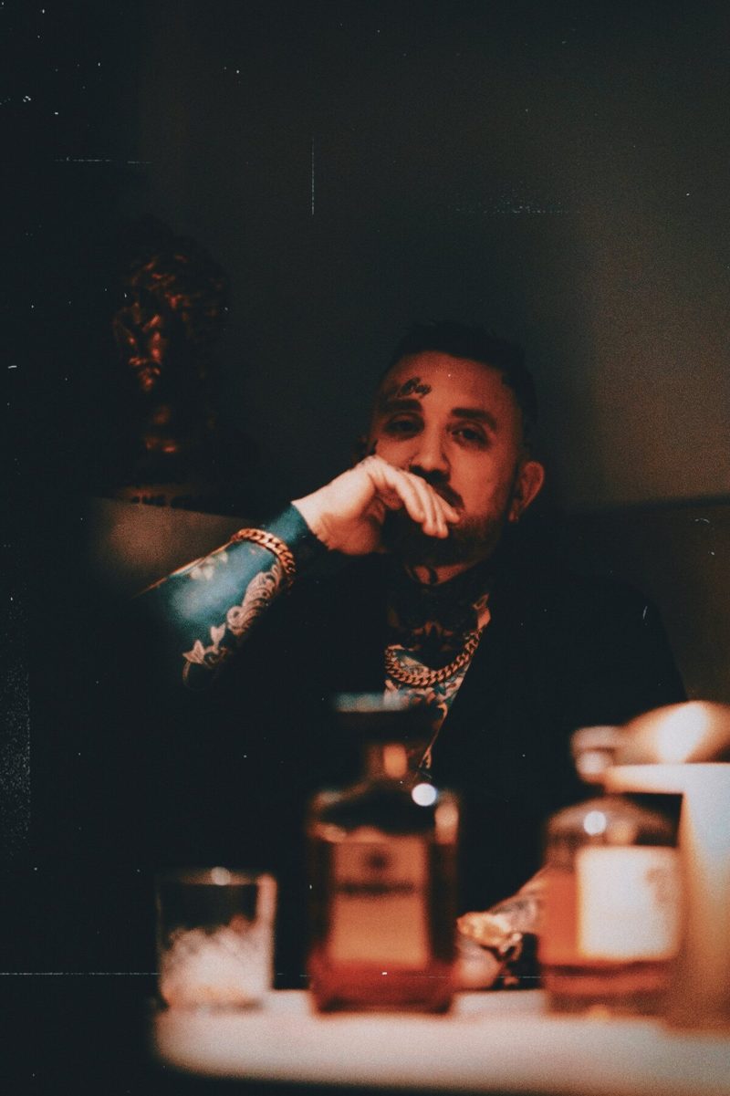 The artist behind Grey Wolf Tattoo, tattooed arms and neck, sitting in low amber light